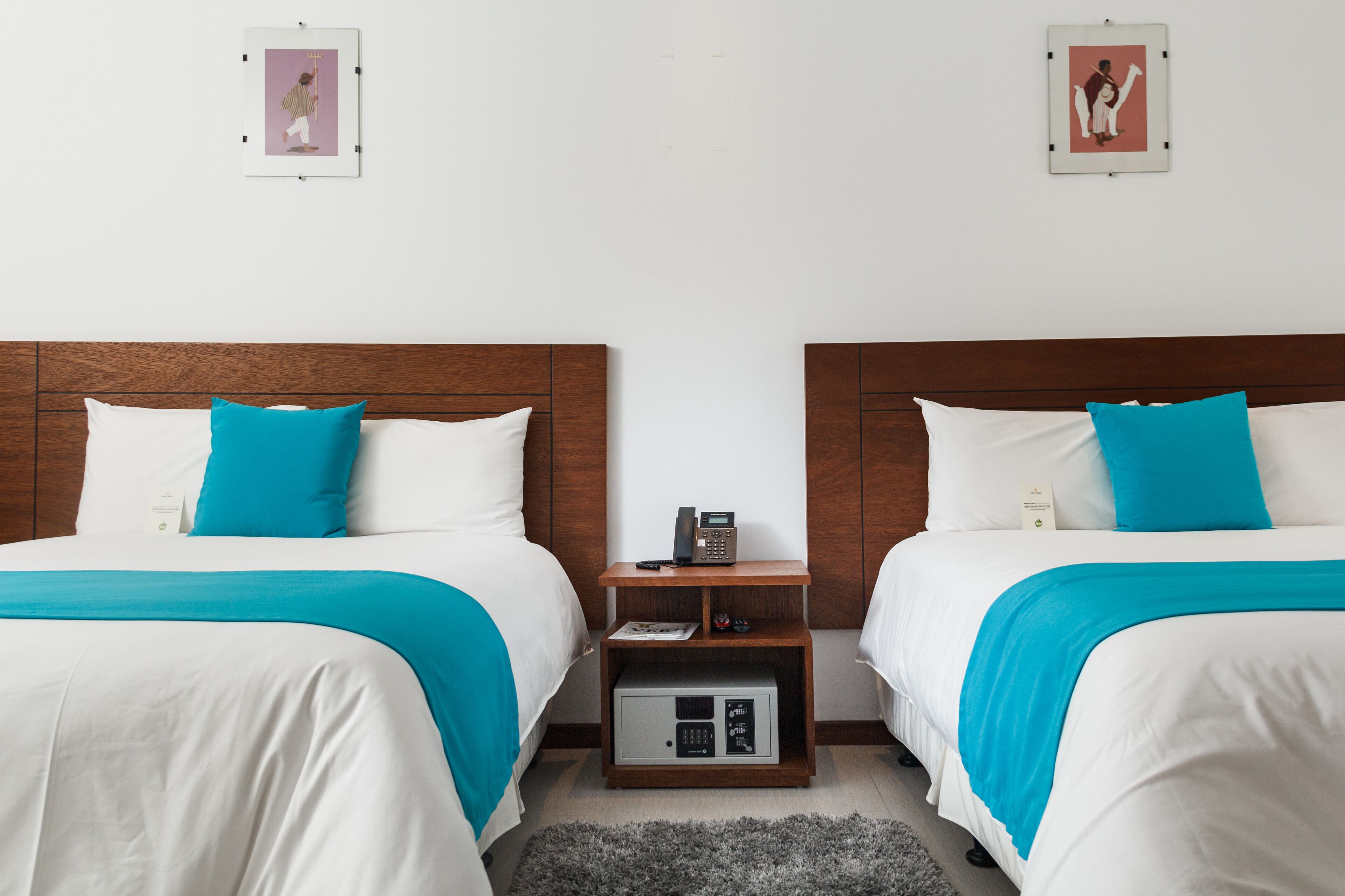 Habitación de hotel con dos camas, cada una con ropa de cama blanca y una manta verde azulada. Hay una mesita de noche de madera entre las camas con un teléfono y un libro pequeño.