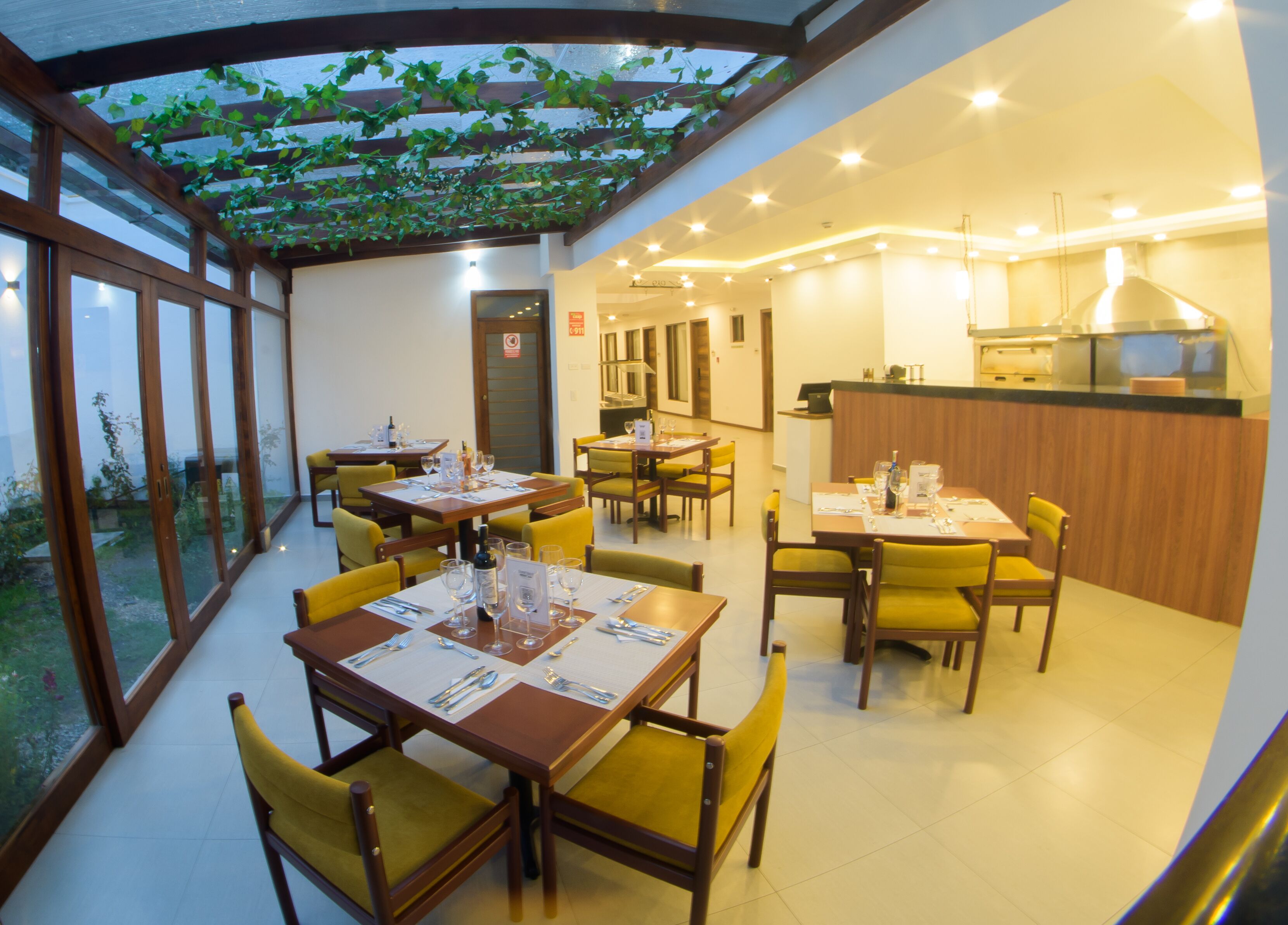 A restaurant with yellow chairs and tables, a kitchen area, and a glass ceiling with greenery.