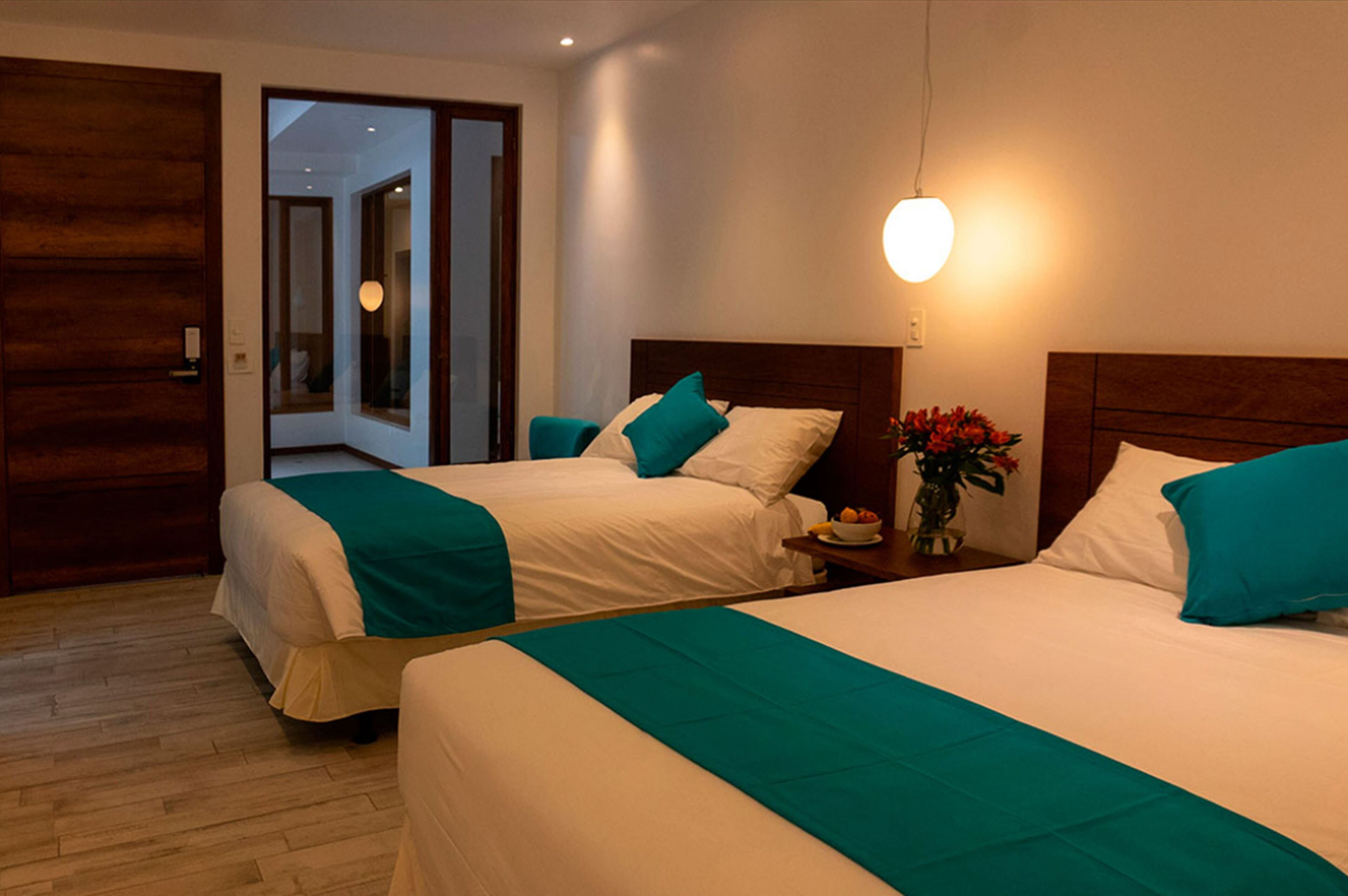 A hotel room with two beds, each with teal bedding, a wooden headboard, and a bedside table with a vase of flowers.