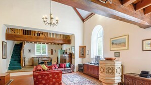 Uma sala de estar ampla, com teto alto, um candelabro, lareira e vista para uma escadaria que dá para outro cômodo.