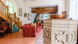 Ampla sala de estar com escada, sofá vermelho estampado, mesa de centro de madeira e um pilar de pedra decorativo.