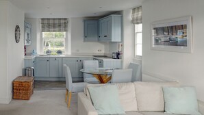 Een moderne keuken met lichtblauwe kastjes, een eethoek met een glazen tafel en stoelen, en een woonkamer met een witte bank en kussens.