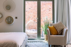 A bedroom with a bed, a chair, a hanging chair, and a view of a brick wall through a glass door.