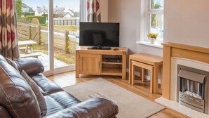 A living room with a leather sofa, a TV on a wooden stand, a fireplace, and a view of the outside. - Gatekeepers Cottage - UK6728 (Lydstep, near Tenby)