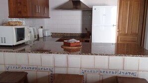 A kitchen with wooden cabinets, a microwave, a toaster, a refrigerator, and a sink. There are also two stools and a blue patterned sofa in the foreground. - Apartamentos en plena naturaleza El Elanio Azul. (Arcos de la Frontera)