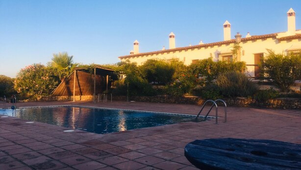 A swimming pool with a stone deck, a covered seating area, and a building with multiple chimneys in the background. - Apartamentos en plena naturaleza El Elanio Azul. (Arcos de la Frontera)