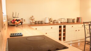 A kitchen with a black stove, wooden cabinets, and a countertop with various appliances and a fruit bowl. - Linley Lane Cottage (Bishops Castle)