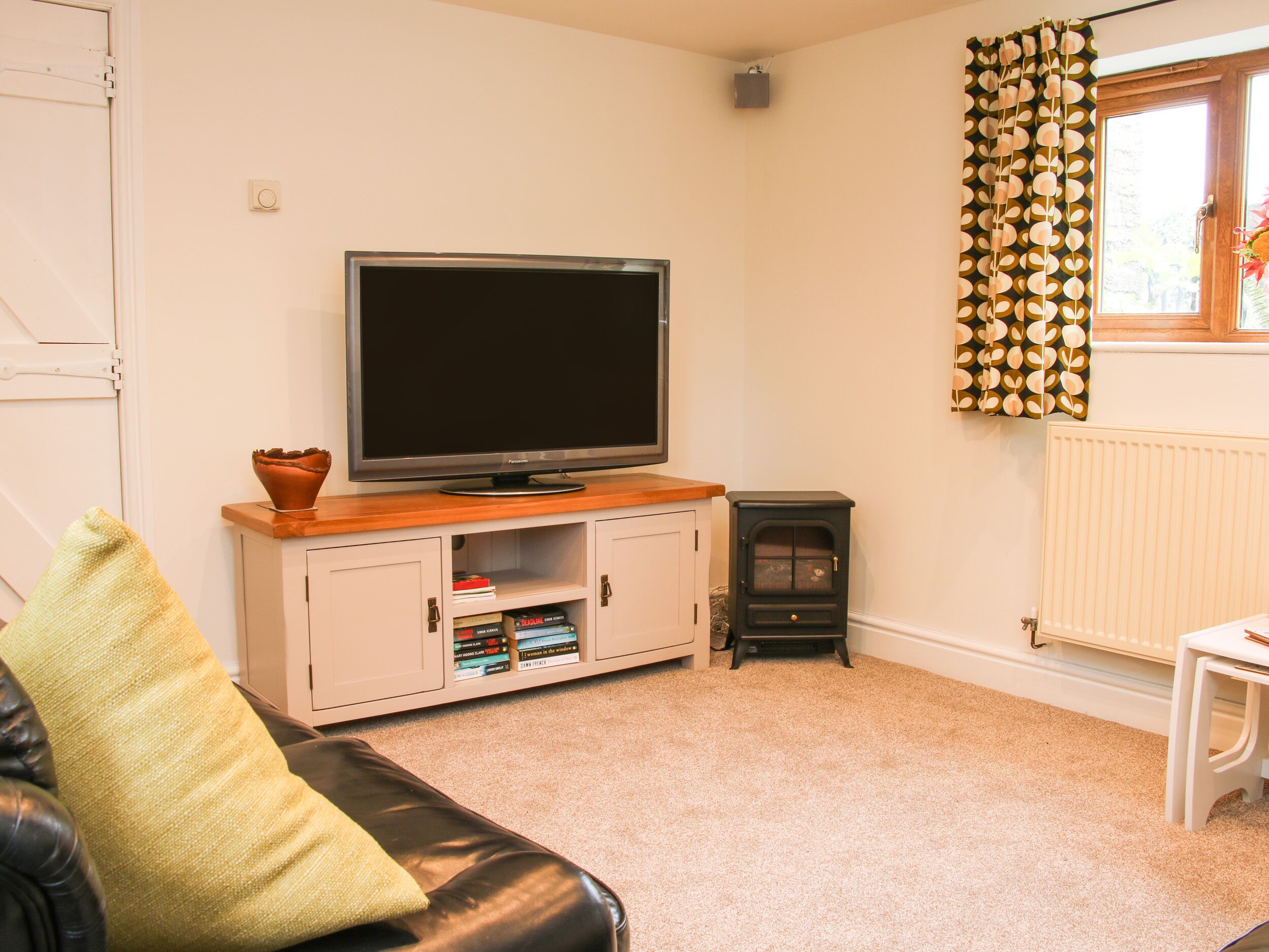 A living room with a television on a wooden stand, a white cabinet with books, a black leather sofa, and a black wood-burning stove.