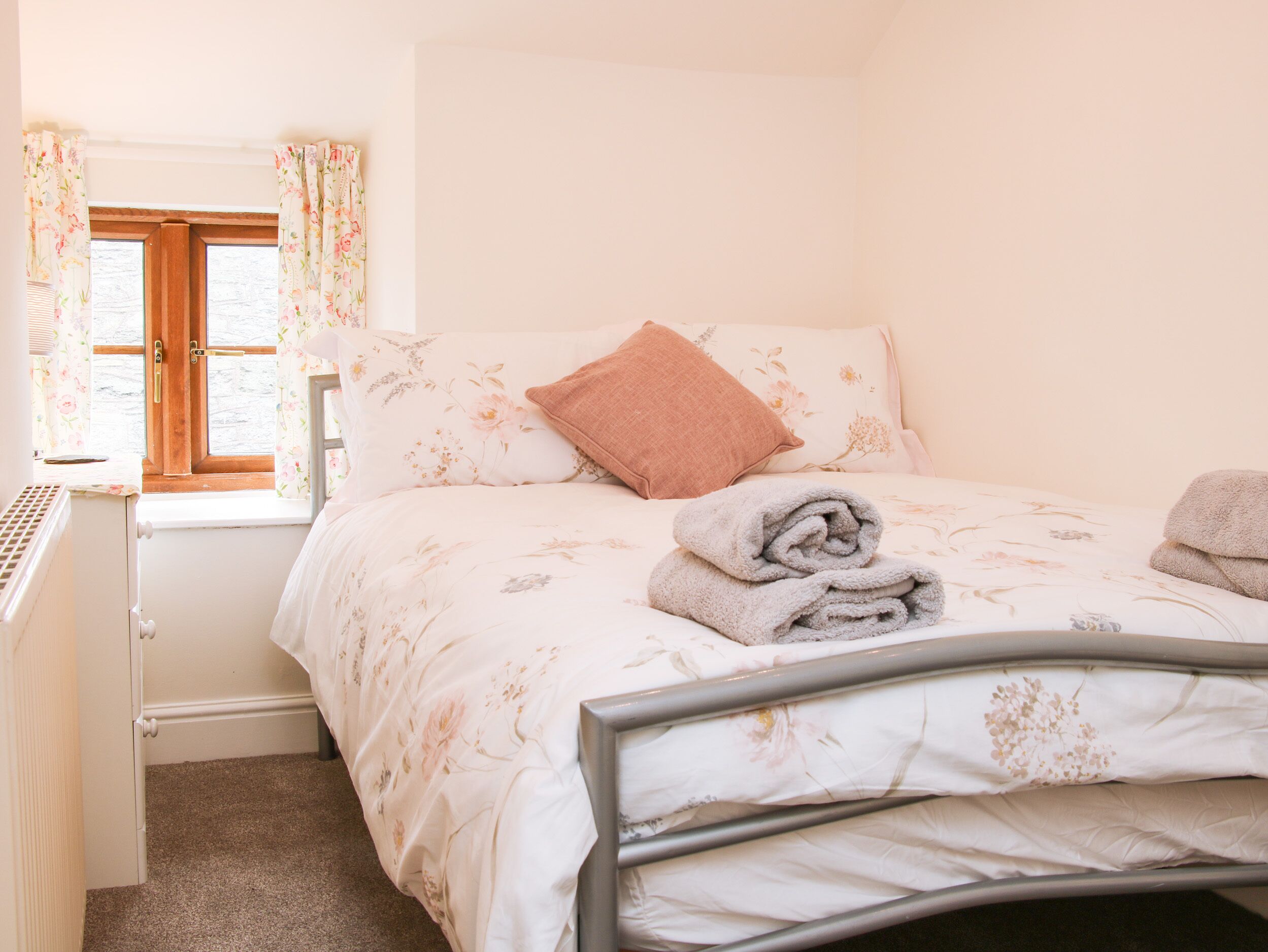 A neatly made bed with floral bedding, a single pink pillow, and rolled towels on the side.