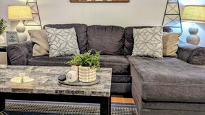 Una sala de estar con un sofá gris oscuro, almohadones decorativos, una mesa de centro con una planta y un letrero, y dos molinos de viento en la pared.