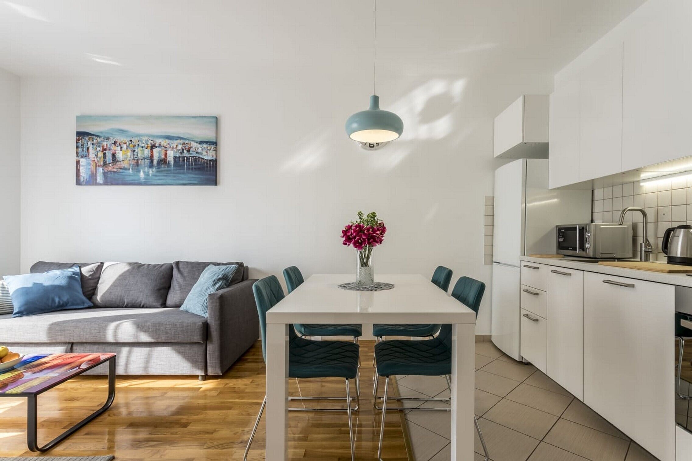 Un salon moderne avec un canapé gris, une table à manger blanche et des chaises turquoise. Un tableau est accroché au mur, et un vase rempli de fleurs est posé sur la table.