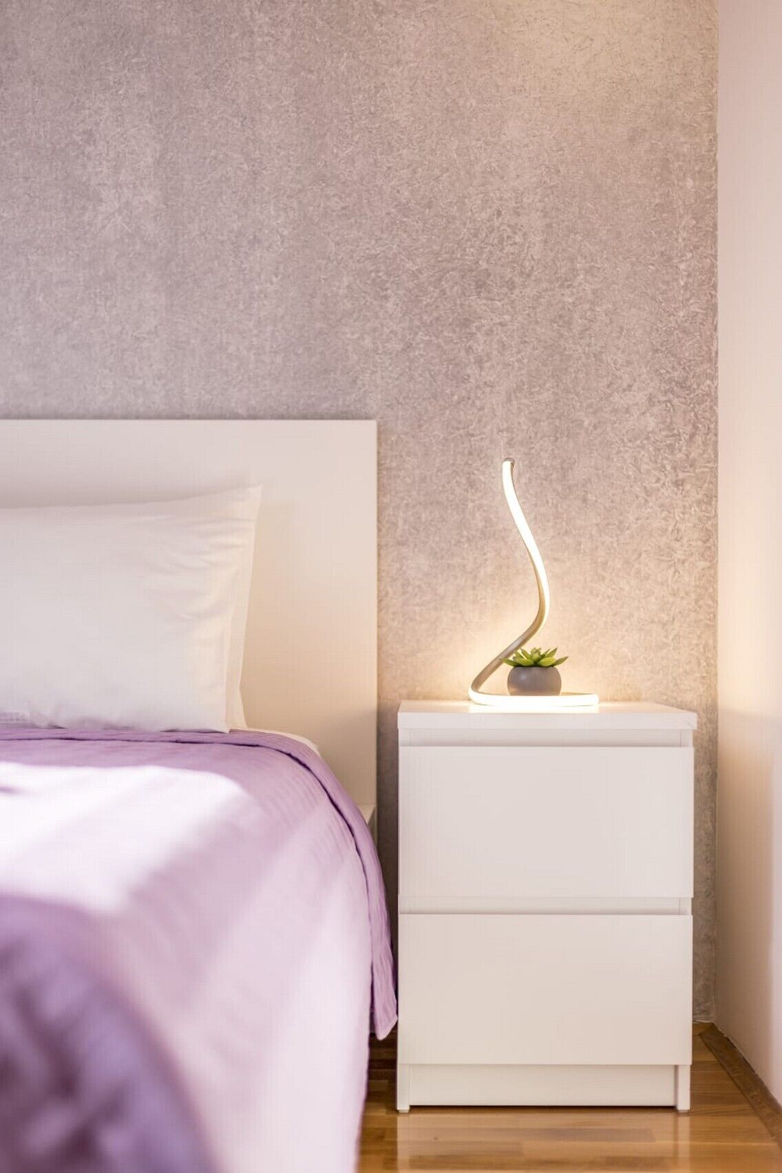 Ein Schlafzimmer mit einem Bett, einem weißen Nachttisch mit Lampe und einer strukturierten Wand.