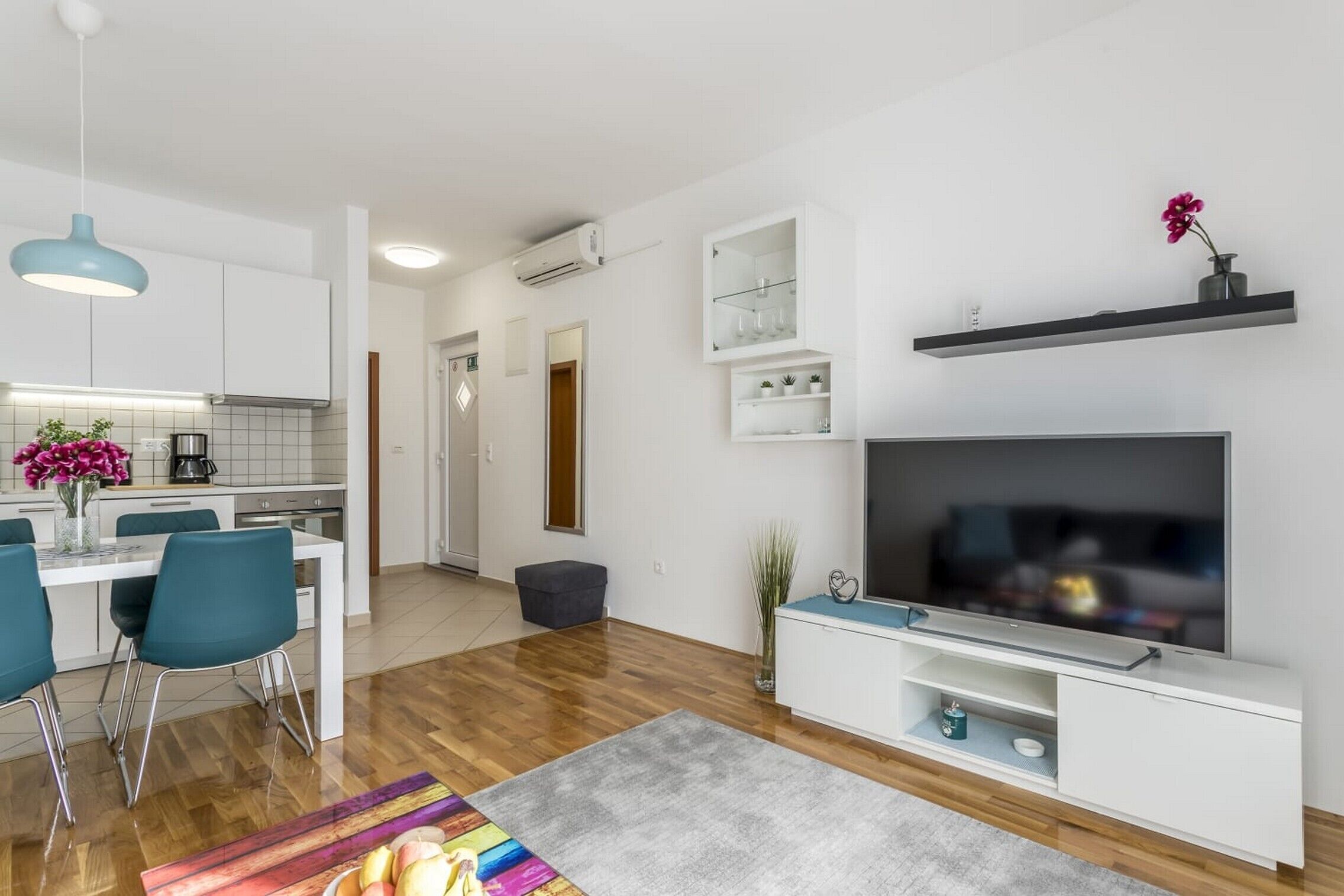 A modern living room with a flat-screen TV, a dining area with chairs and a table, and a kitchen with white cabinets and a blue pendant light.