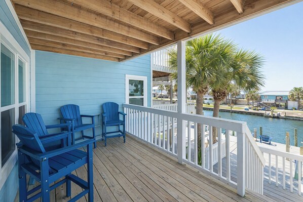 Une terrasse en bois avec des chaises Adirondack bleues, une balustrade blanche et une vue sur une marina avec des bateaux et des palmiers.