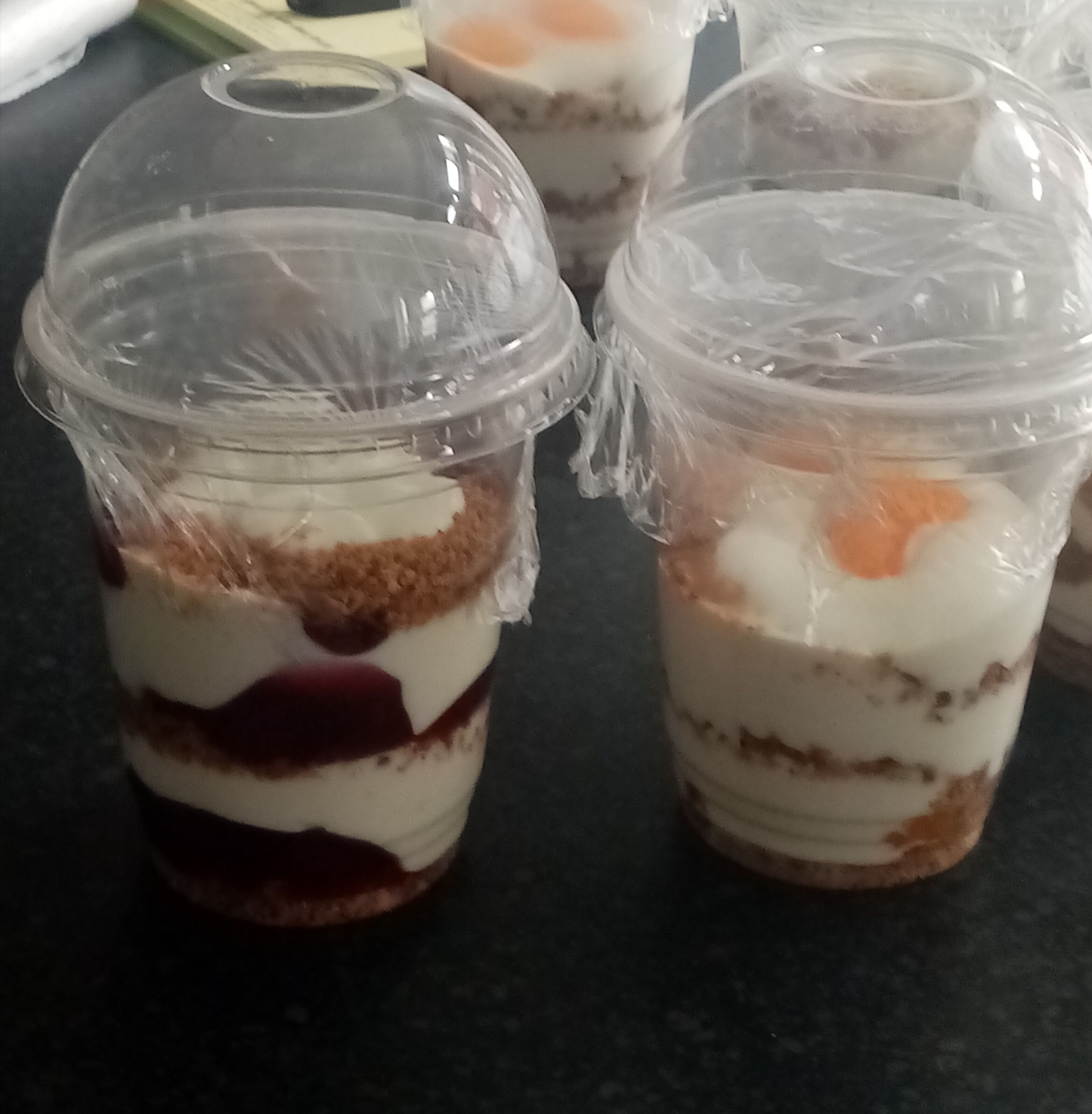 Three layered dessert cups with a clear plastic lid, placed on a dark countertop.