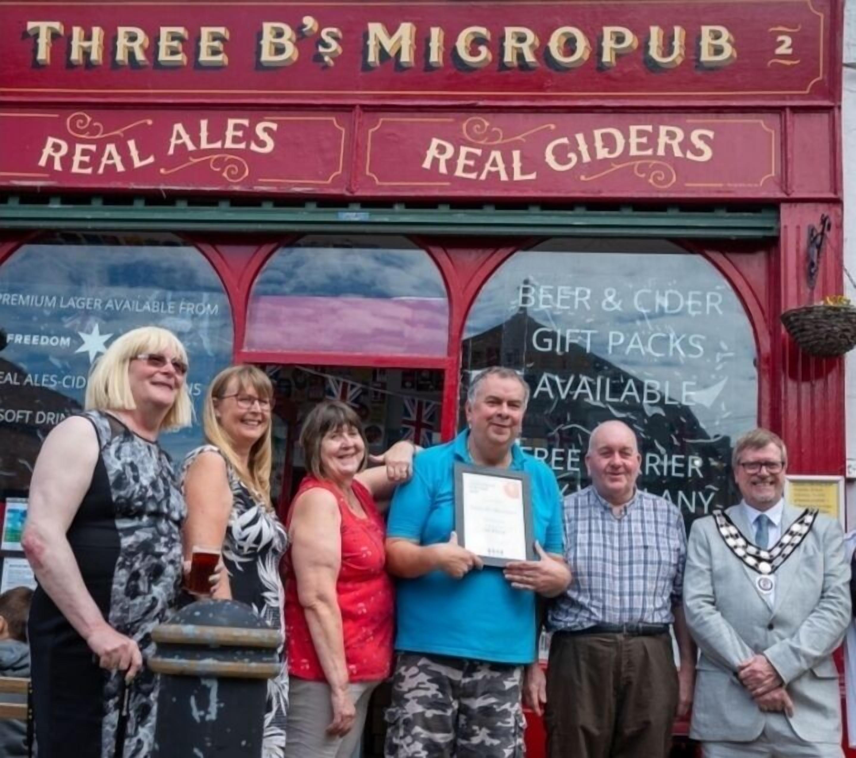 Un groupe de personnes se tient devant un pub appelé « Three B’s Micropub 2 », avec des panneaux publicitaires pour des « bières artisanales » et des « cidres artisanaux ».