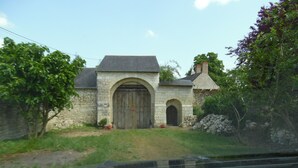 Un bâtiment en pierre doté d’une grande porte en bois et d’une porte plus petite, entouré de verdure et délimité par un mur de pierre.