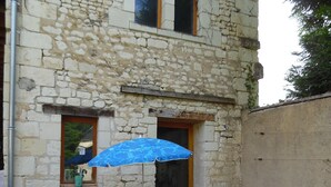 Il s’agit d’un bâtiment en pierre doté d’un petit patio. On y trouve une table entourée de chaises et un parasol bleu. Une chaise sur laquelle est posée une serviette à rayures est également visible.
