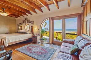 A bedroom with a bed, a wooden ceiling, a large window with a view of the sea, a brown leather sofa, and a patterned rug. - Oceanfront Master Suite (St. Augustine)