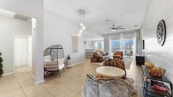 A spacious living area with a high ceiling, a ceiling fan, and a large window providing a view of the outdoors. There is a seating area with a round table and chairs, and a decorative wall clock.