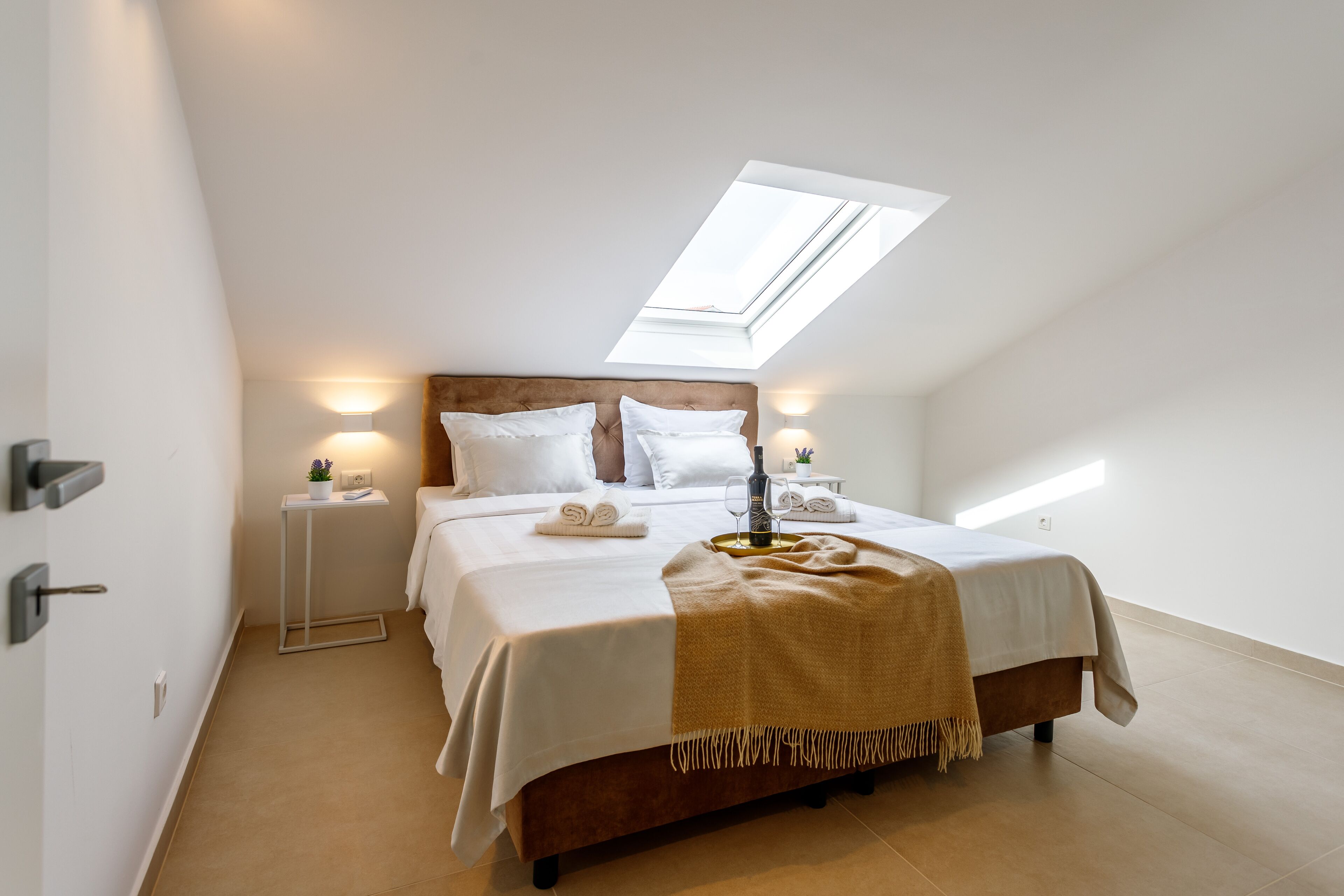 A bedroom with a skylight, a bed with white linens, a bedside table with a lamp, and a small plant.