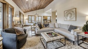 A spacious living room with a wooden ceiling, a large sofa set, a dining area, and a kitchen in the background.