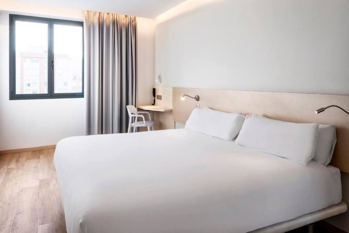 A hotel room with a large bed, a desk, a chair, a window with curtains, and a headboard with a light fixture.