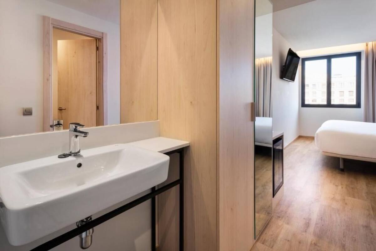 A modern bathroom with a white sink, a wooden door, and a view of a bedroom with a bed and a TV.