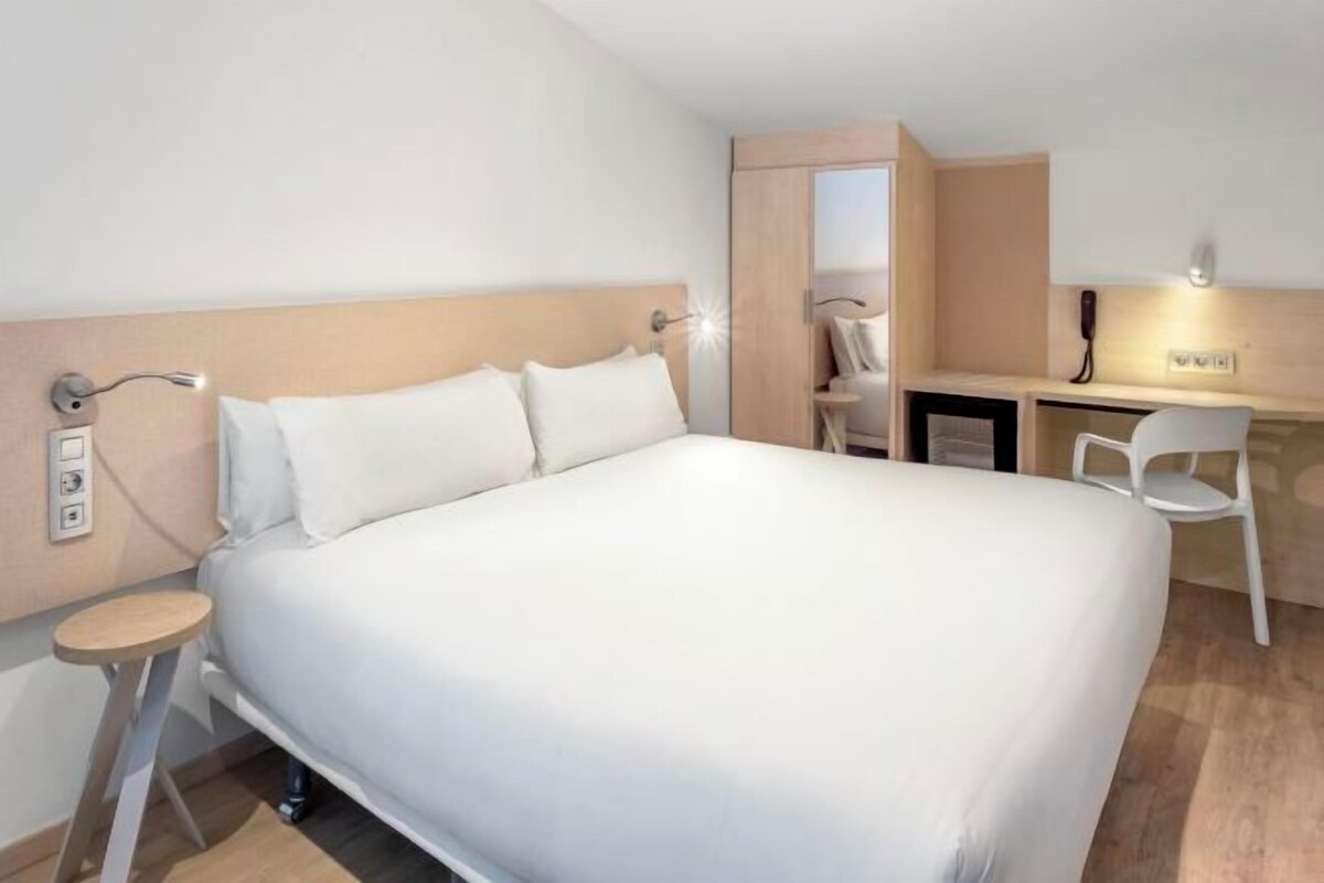A hotel room with a large bed, a desk with a chair, a telephone, and a mirror.