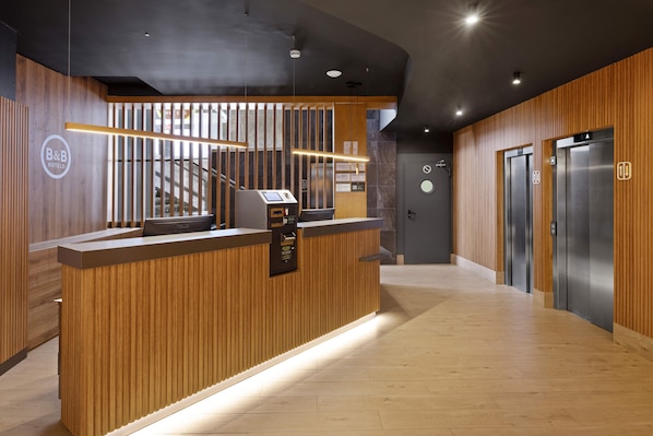A reception desk with a wooden finish, a digital check-in kiosk, and two elevators. - B&B Hotel Málaga Centro (Málaga)