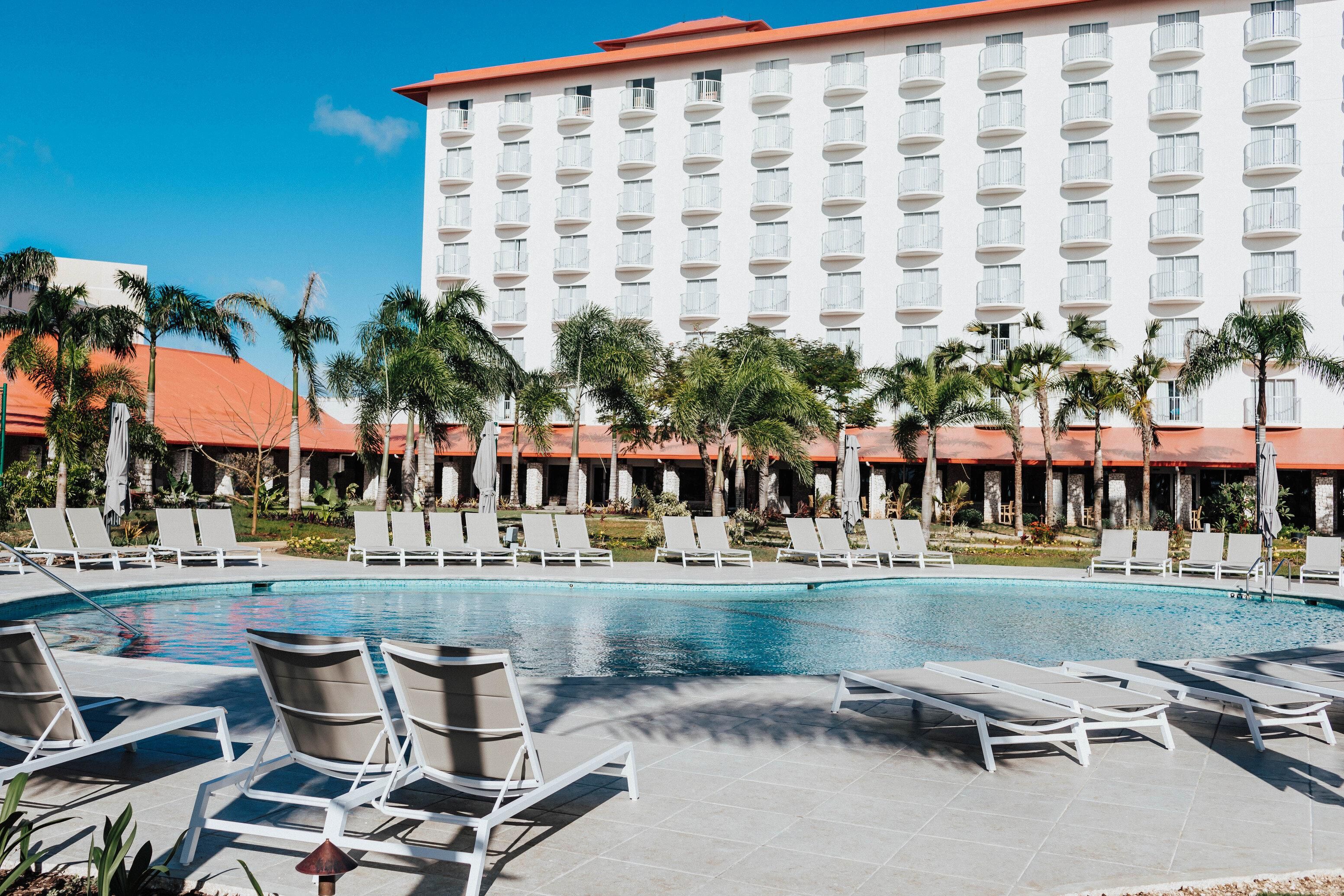 A hotel with a swimming pool, lounge chairs, palm trees, and a large building with balconies.
