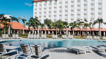 A hotel with a swimming pool, lounge chairs, palm trees, and a large building with balconies.