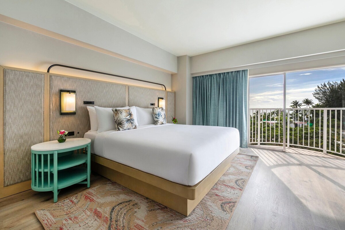 A hotel room with a large bed, a small side table, a nightstand, a large window with a view, and a balcony.