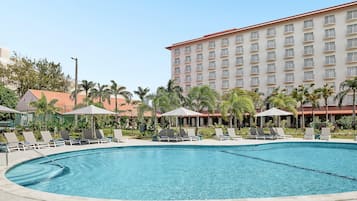 Una piscina con sillas de descanso y sombrillas, rodeada de palmeras y un edificio de hotel.