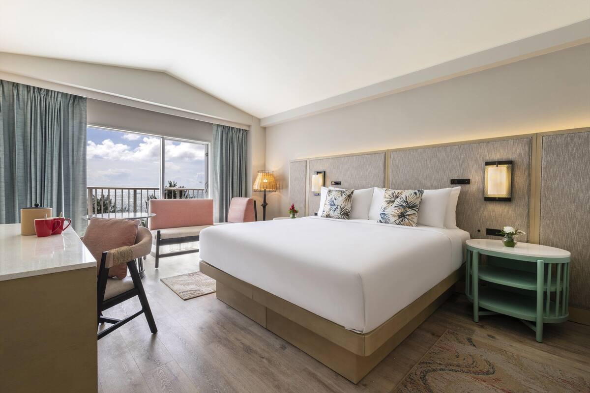 A hotel room with a large bed, a desk, a chair, a nightstand, and a view of the outdoors.