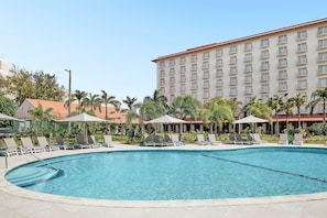 A swimming pool with lounge chairs and umbrellas, surrounded by palm trees and a hotel building.