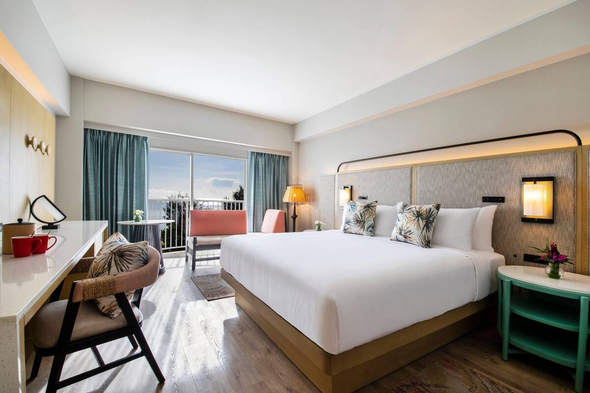 A hotel room with a large bed, a desk with a chair, a small table, and a balcony with a view.