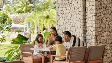 A family dining at an outdoor table with wicker chairs, a stone wall, and lush greenery in the background.