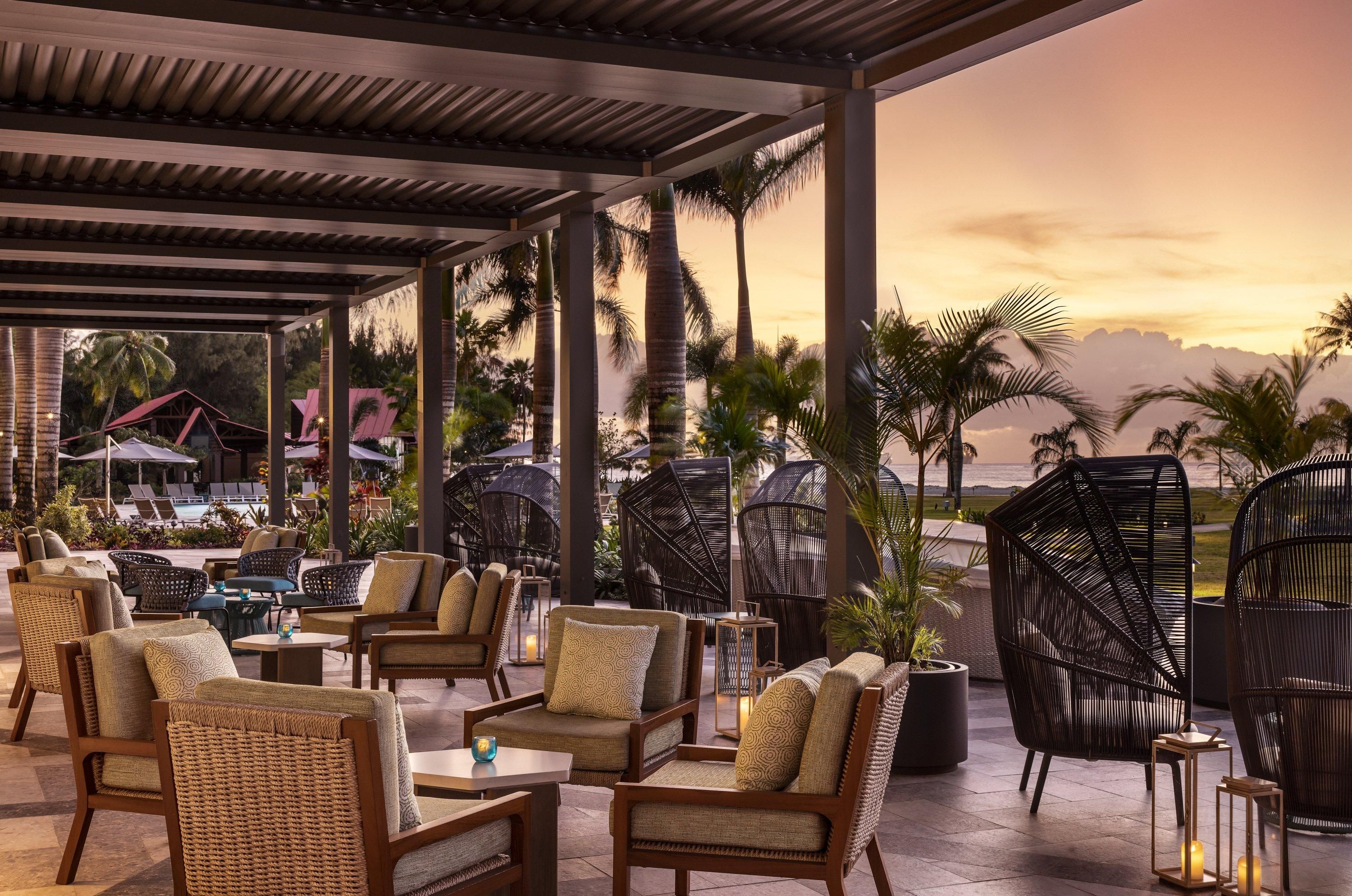 A covered outdoor seating area with wicker furniture, palm trees, and a view of the ocean at sunset.
