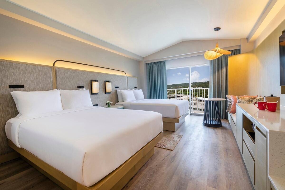 A hotel room with two beds, a kitchenette, a balcony with a view, and a hanging light fixture.