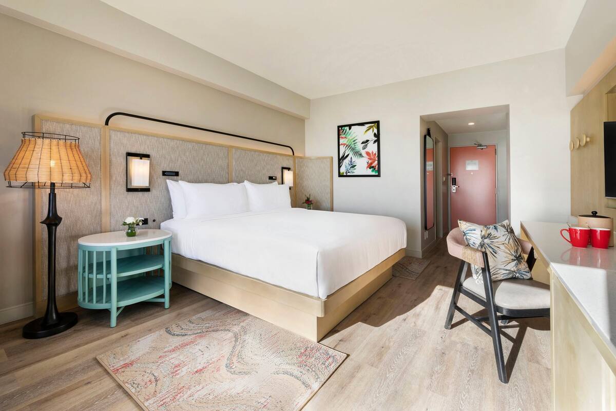 A modern hotel room with a bed, bedside lamp, small table, chair, and a view of the hallway.