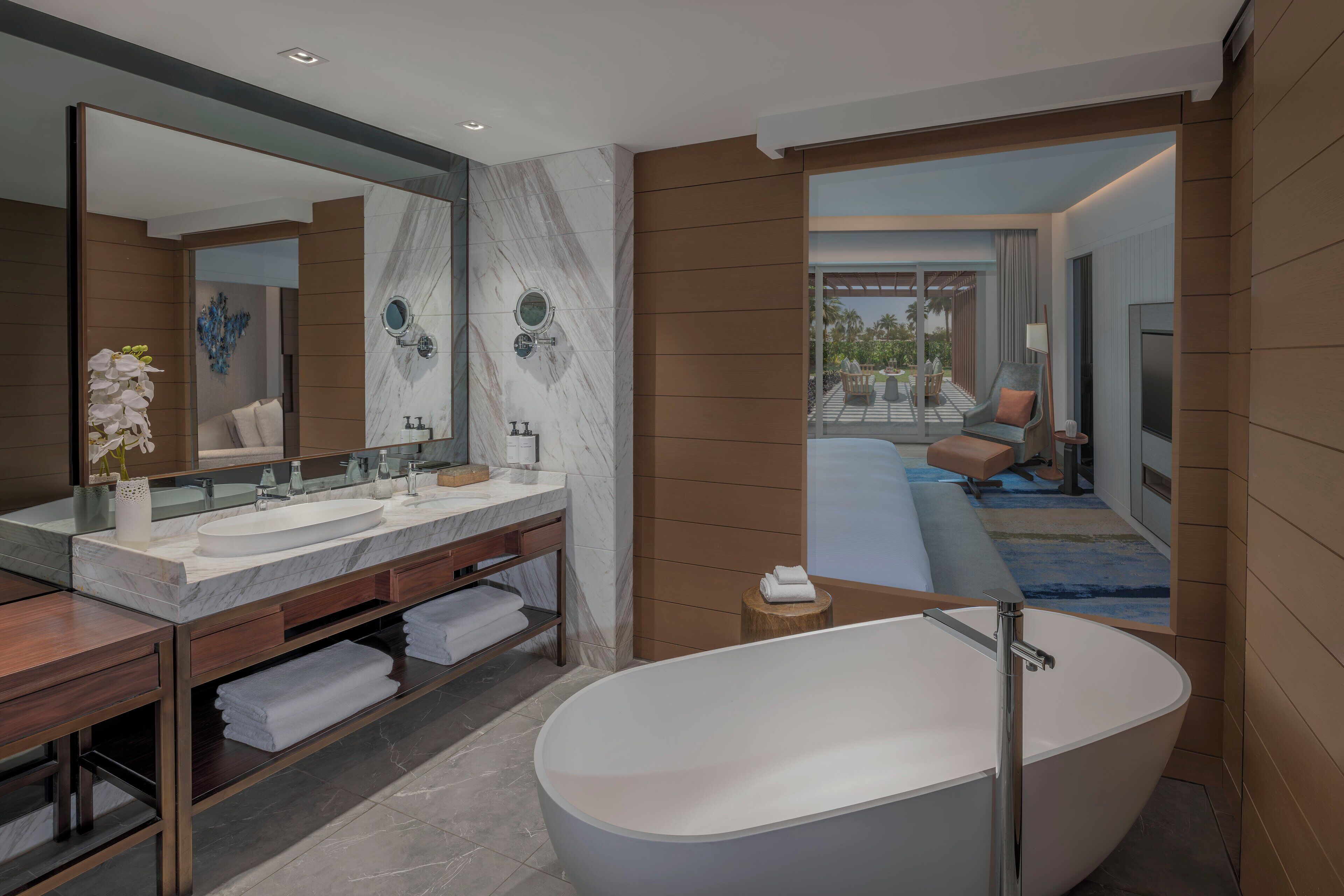 A modern bathroom with a freestanding bathtub, a large vanity with a mirror, and a view of a balcony with outdoor seating.