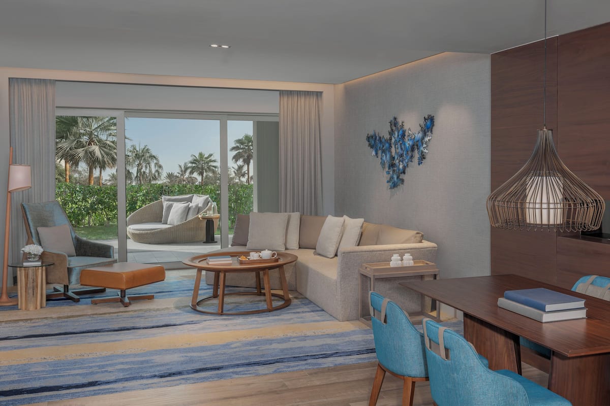 Una habitación de hotel moderna con sofá, una mesa de comedor con sillas, una mesa de centro y vistas a palmeras a través de amplios ventanales.