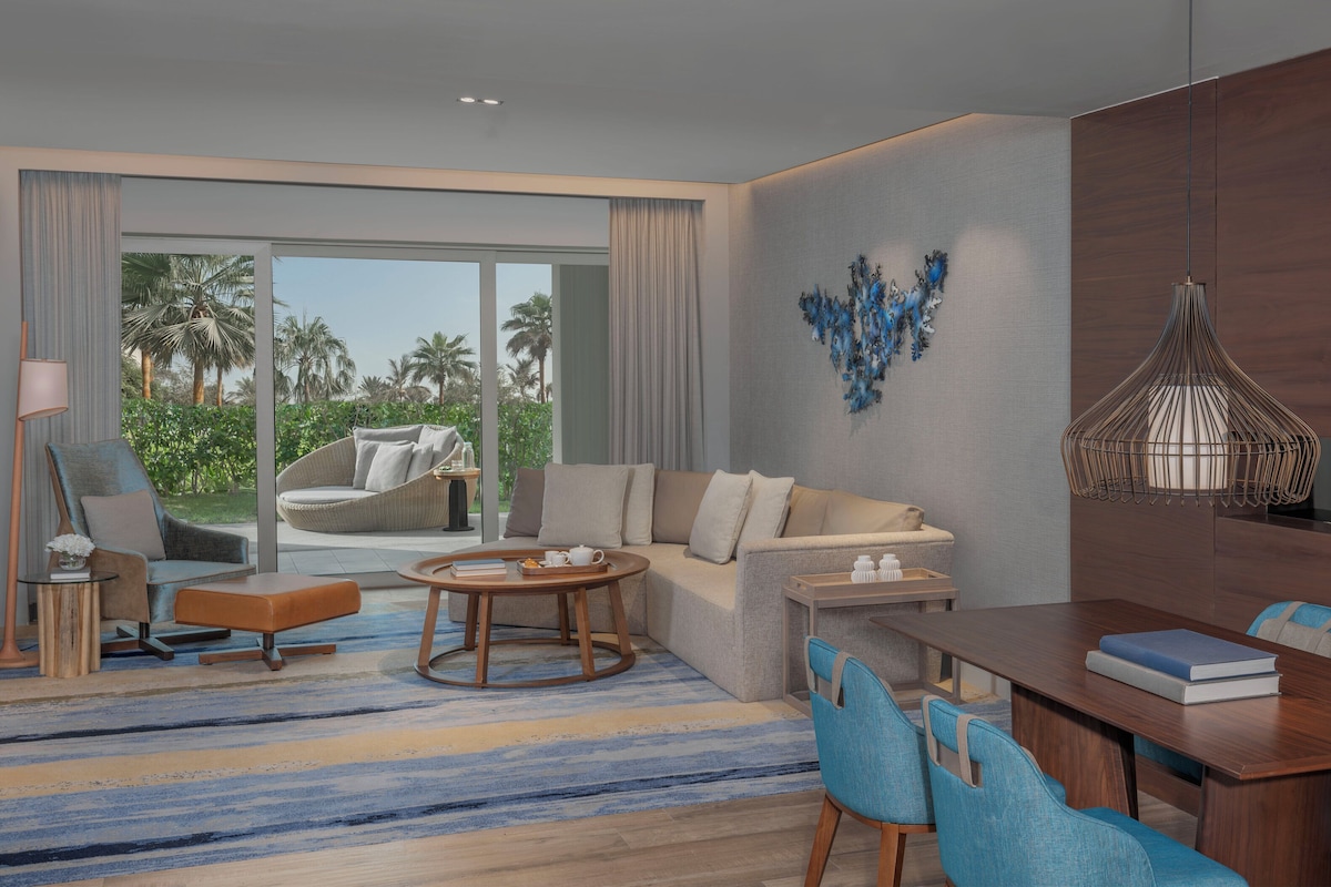A modern hotel room with a sofa, a dining table with chairs, a coffee table, and a view of palm trees through large windows.