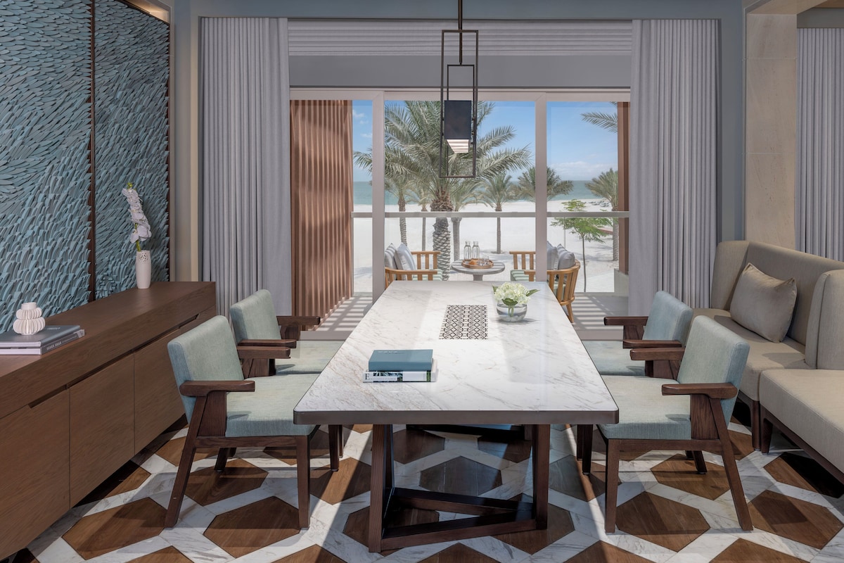A modern dining area with a marble table, wooden chairs, and a view of the beach.