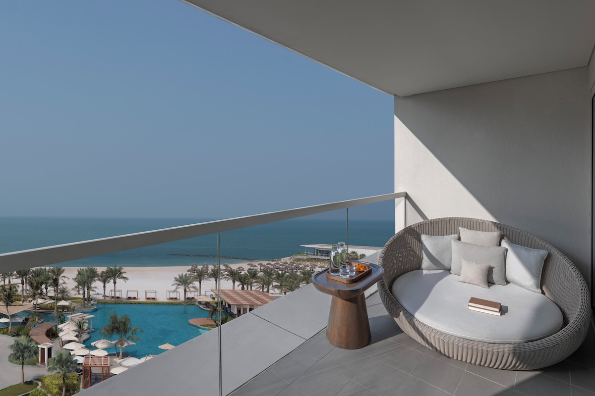 A balcony with a wicker sofa, a small wooden table with drinks, and a view of a beach and pool.