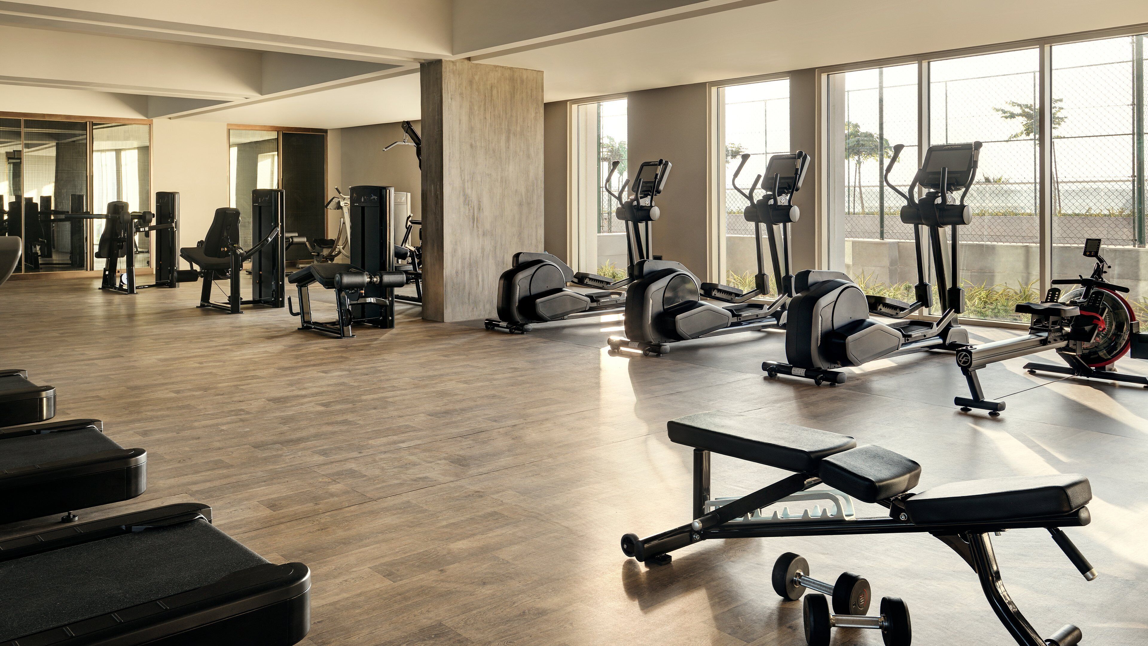 A spacious gym with various exercise equipment, large windows, and a wooden floor.