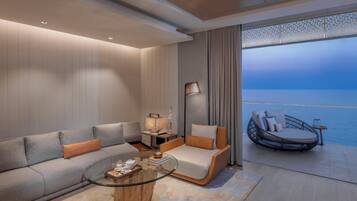 A modern hotel room with a sofa, a glass coffee table, a daybed, a lamp, and a view of the sea.