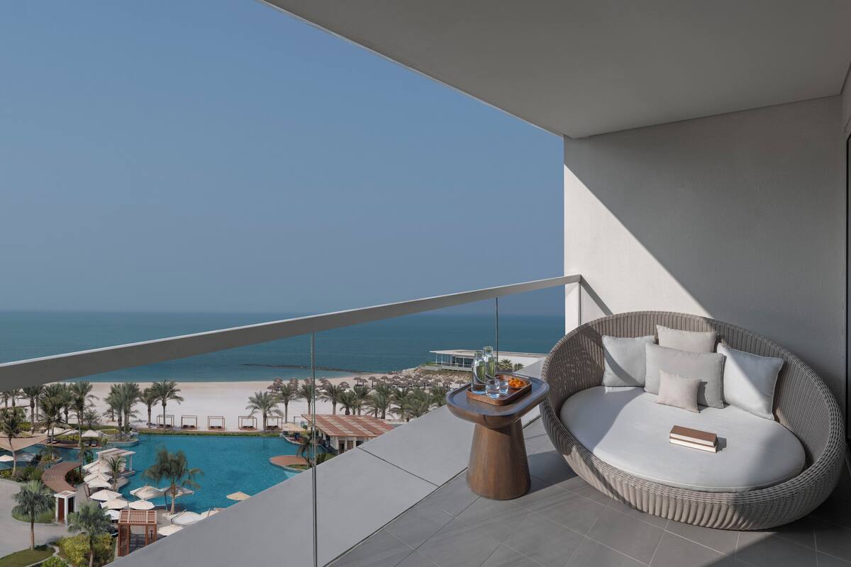 A balcony with a wicker sofa, a small wooden table with drinks, and a view of a beach and pool.
