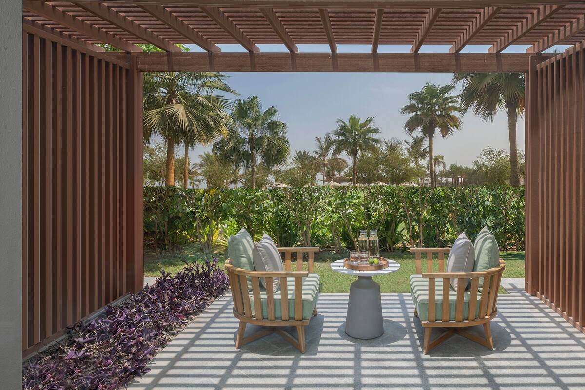 A covered outdoor seating area with two wooden chairs and a table, surrounded by lush greenery and palm trees.